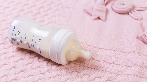 Image of a baby bottle half-filled with milk placed on a bed with a pink bedsheet.