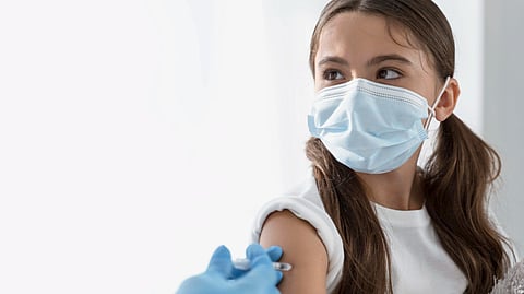 An image of a girl in a mask wearing a white t-shirt while getting vaccinated.