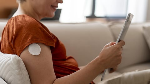 An image of a woman in a rust-colored shirt siting on a beige sofa, a medical patch is visible on her upper arm.