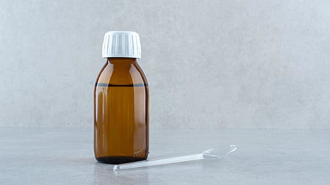 Image of a bottle of cough syrup along with a plastic spoon on a white surface.