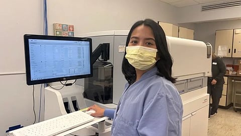 An image of a laboratory technician wearing a yellow face mask and blue lab coat stands beside a large machine looking at the camera. 