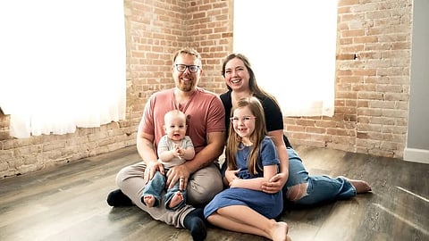 An image of a smiling family of four sits on a wooden floor against a brick wall.