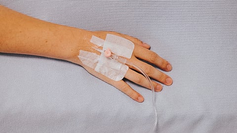 An image of a patient's hand with an IV drip attached, resting on a hospital bed.