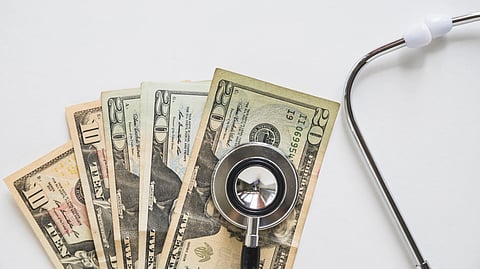 An image of stethoscope rests on fanned-out U.S. dollar bills, including tens and twenties, against a white background.