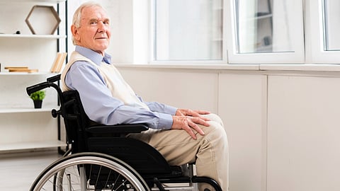 An image of elderly man in a wheelchair sits near a bright window, wearing a light blue shirt and beige vest.