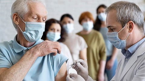 An elderly man wearing a mask receives a vaccine shot from a healthcare worker in a white coat.