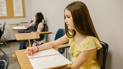 An image of a young girl in a yellow dress focused on writing in a notebook at a classroom desk. 
