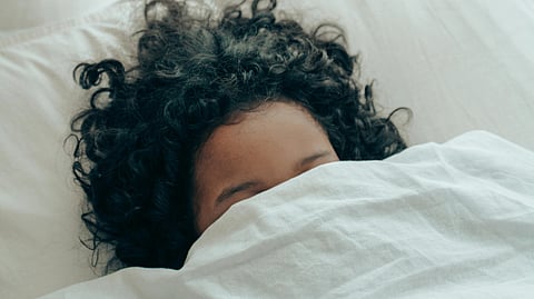 Image of a woman sleeping in bed, covered with a white bedsheet and white blanket up to her face.