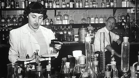An image of a woman in a lab coat measuring liquids on a balance in a cluttered laboratory. 