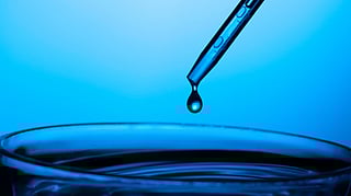 An image of blue-tinted close-up of a single droplet from a pipette about to fall into a glass petri dish.
