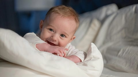 An image of smiling baby with light brown hair in a bed having white pillows and sheet.
