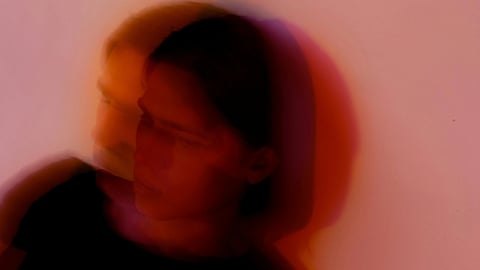 A blurred image of a person in a black shirt against a pink and orange background.