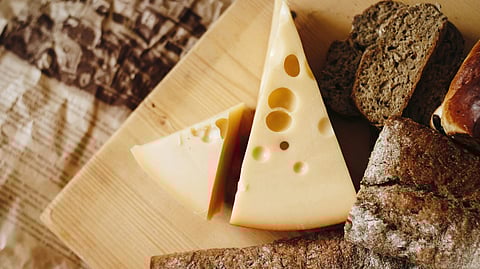 Image of a block of cheese in triangular shape over a wooden board with brown bread and brownie.