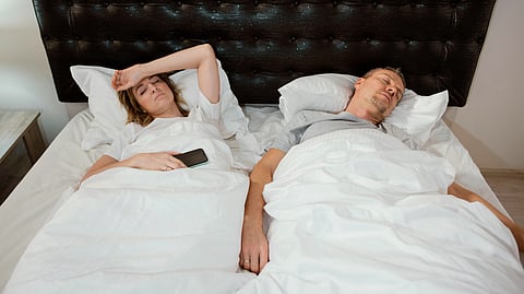 An image of woman lies awake holding a phone, looking frustrated on a bed with white sheets, while a man peacefully sleeps beside her.