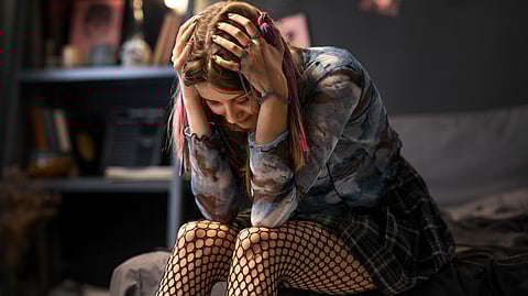 An image of a young woman sits on bed, head in hands, wearing fishnet tights and a patterned top.