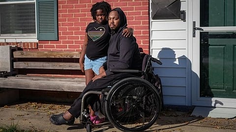 An image of man in a wheelchair sits beside a young girl outside a brick building.