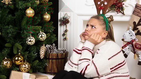 An image of a woman with reindeer antlers sits by a Christmas tree.