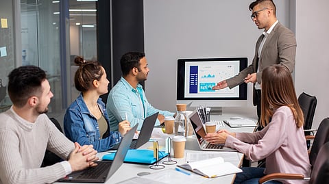 An image of a man in a suit presenting graphs on a screen to colleagues seated with laptops in a conference room.
