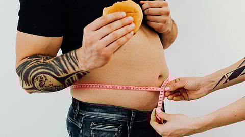 An image of tattooed man in a black shirt holds a burger while another person measures his waist with a pink tape.