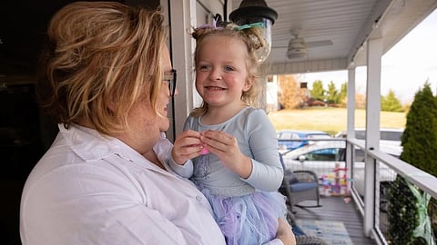 An image of smiling child in a pastel tutu dress is held by an adult on a porch. They share a joyful moment.