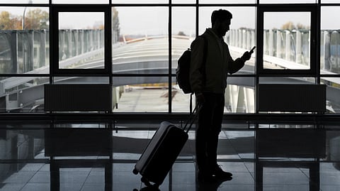 An image of a man with a suitcase and backpack checking his phone at an airport terminal.