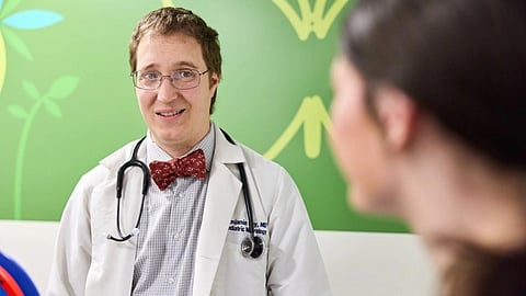 An image of doctor in a white coat with a stethoscope and a red bow tie smiles kindly at a patient.