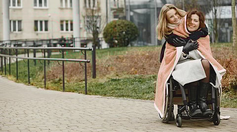 An image of a woman sitting in wheelchair and another woman hugging her from behind.