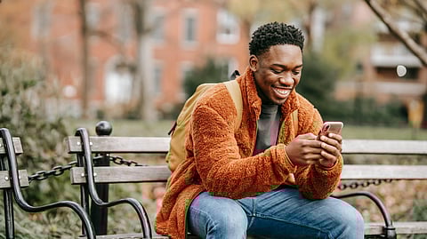 An image of man in a brown fleece jacket sits on a park bench, smiling at his phone.