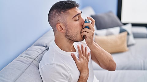 An image of a man in a white shirt using a blue inhaler on a light gray sofa, hand on his chest. 