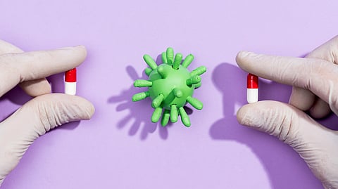 An image of gloved hands hold red and white capsules beside a green virus model on a purple background