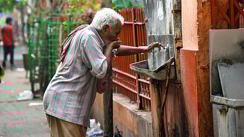 An elderly man with gray hair drinks from a public water tap attached to a weathered wall on a street. 