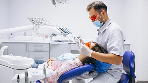 An image of a dentist wearing protective glasses and a mask works on a patient reclined in a dental chair.