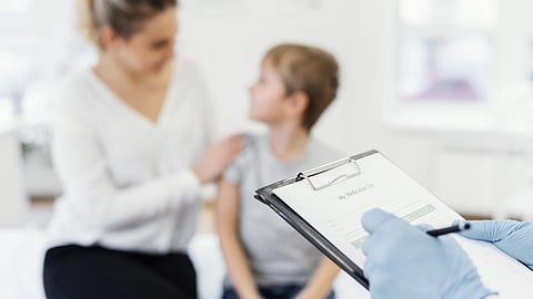 A mother and a boy child in a hospital bed and a doctor taking notes about the patient on chart.