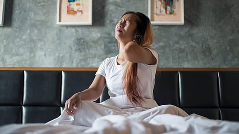 An image of a woman in a white shirt sits on a bed and holding her neck in pain.