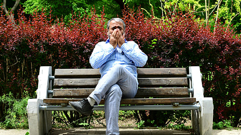An image of a man in a blue shirt and gray pants sits on a bench in a park. His hands cover his face,surrounded by red foliage.