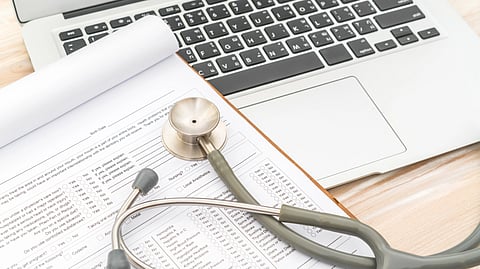 An image of stethoscope resting on a medical form next to an open laptop on a wooden desk.