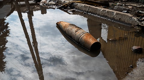 An image of dusty, cylindrical object lies in a puddle reflecting sky and clouds, near littered pavement. 