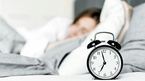A woman peacefully sleeps in bed beside an alarm clock displaying the time