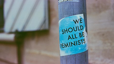 An image of street pole displays a blue and white sticker reading "We Should All Be Feminists." 
