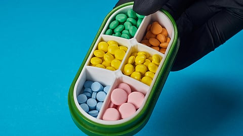 An image of gloved hand holds a pill organizer with colorful compartments against a blue background.