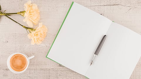 An open notebook with a green edge lies on a light wooden table. A gray pen rests on its pages. Nearby, a cup of coffee and two flowers.