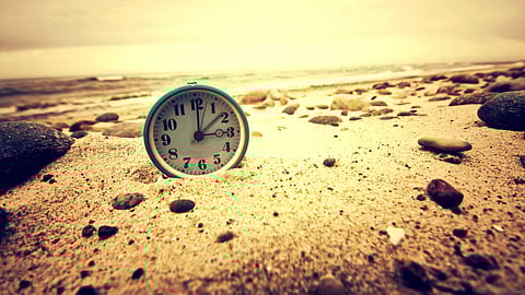 An image of vintage clock half-buried in beach sand, surrounded by small pebbles.