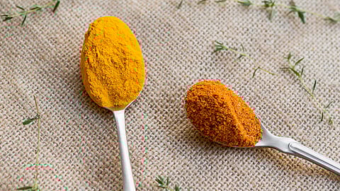 An image of two metal spoons on burlap, one with vibrant yellow turmeric and the other with reddish-orange.
