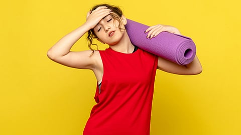 An image of a woman in a red tank top stands against a bright yellow background, eyes closed, hand on forehead, and a purple yoga mat on her shoulder.