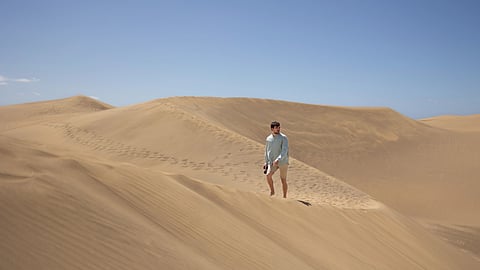 An image of a person walks on vast, sunlit sand dunes under a clear blue sky. 