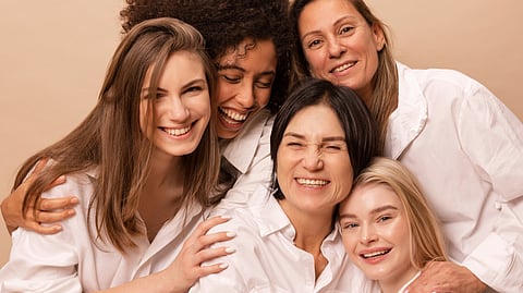 An image of five women in white shirts joyfully embrace against a beige background.