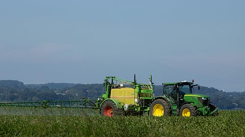 An image of green tractor with a large yellow tank attachment is spraying a vast green field under a clear blue sky.