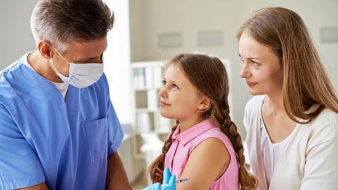 An image of a nurse in blue scrubs, wearing a mask, administers a vaccine to a young girl with braids, sitting beside her smiling mother.