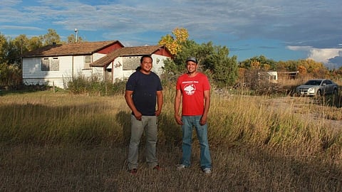 An image of two men stand smiling in front of a rustic white house, surrounded by tall grass and trees.