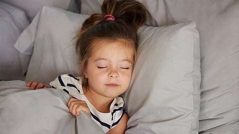 An image of a young girl with a ponytail sleeping peacefully on a bed with gray pillows and sheets.